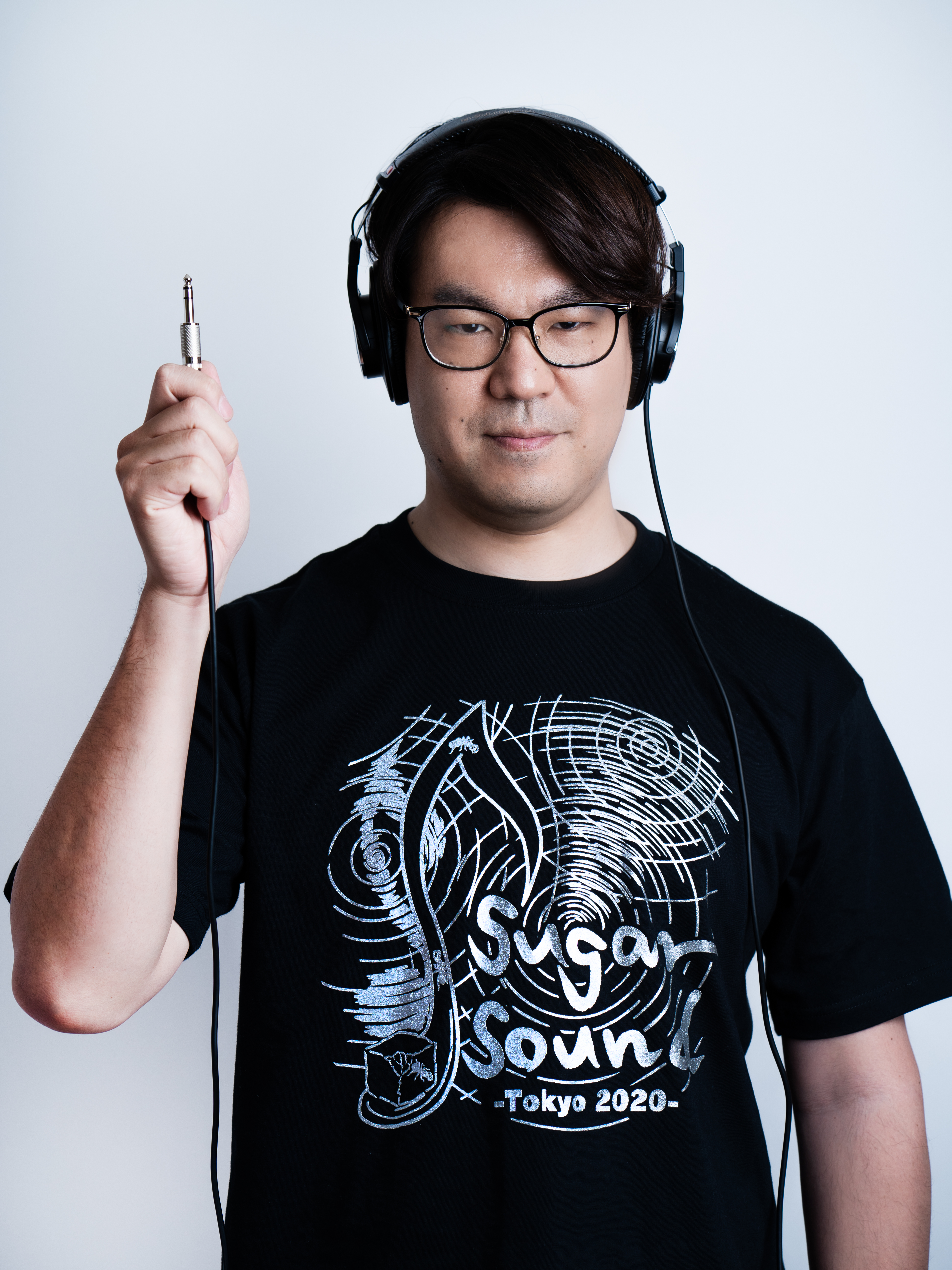 佐藤こうじさんのお写真。「Sugar Sound」というロゴの入った黒いTシャツを着て、業務用ヘッドフォンをつけ、右手でそのプラグを持ってその手を挙げている